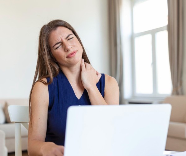A woman sitting in front of a laptop, holding the side of her neck with a look of pain on her face.